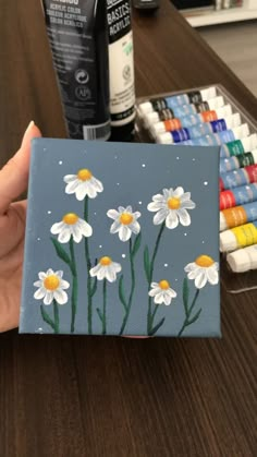 a person is holding up a painted canvas with daisies on it and crayons in the background