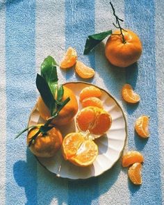 oranges are on a plate with leaves and peeled ones next to them, sitting on a blue striped tablecloth