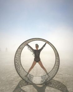 a woman standing in front of a large metal object on the beach with her arms outstretched