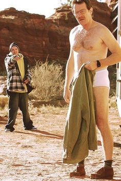 a man with no shirt standing in the desert next to another man holding a towel