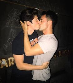 two people are kissing each other in front of a wall with writing on it and one person has his arm around the woman's neck