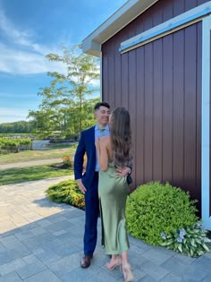 a man and woman standing next to each other in front of a brown building with green plants