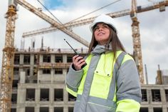 a woman in safety gear standing next to a construction site holding a cell phone and looking up at the sky