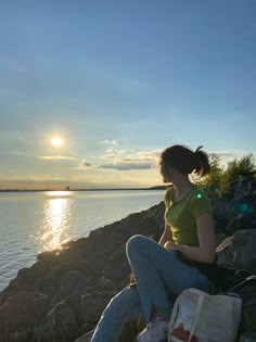 a woman sitting on top of a rock next to the ocean under a sun set