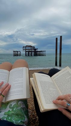 two people are sitting on the beach reading books and looking at the ocean with an old pier in the background