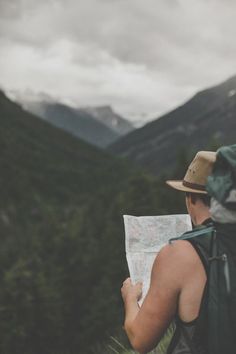 a man with a hat and backpack looking at a map in the mountains on a cloudy day