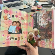 a hand holding up a scrapbook with two pictures on the cover and an open laptop in the background