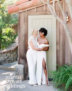 two women standing next to each other in front of a wooden structure with trees and bushes