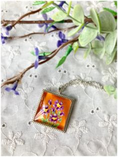 an orange square pendant with purple flowers on it sitting on a white lace tablecloth