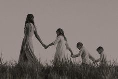 three children holding hands while walking through tall grass in black and white photo with two adults standing behind them