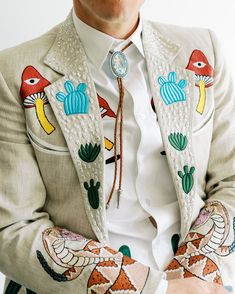 a man in a white shirt and jacket with colorful embroidered patches on his arms, sitting down