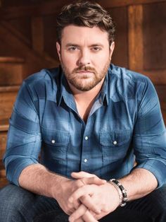 a man sitting in front of a wooden wall wearing a blue shirt and black pants