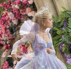 a woman in a blue dress and tiara walking down the street with flowers behind her