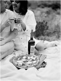 two people sitting on the ground with food and wine in front of them, one holding a bottle