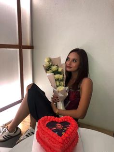 a woman sitting in front of a heart shaped cake with flowers on the table next to it