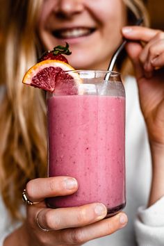 a woman holding a glass with a pink smoothie in it and an orange slice on top