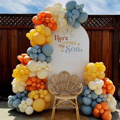 there is a chair and balloon arch in front of a sign that says here comes the son