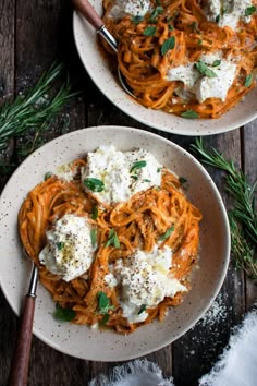 two white bowls filled with pasta and cheese on top of a wooden table next to herbs