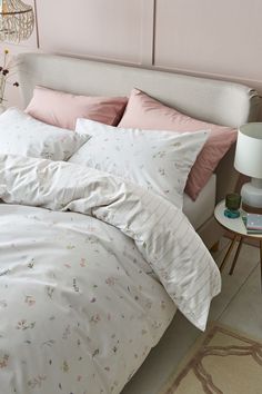 a white bed with pink sheets and pillows in a room next to a lamp on a table