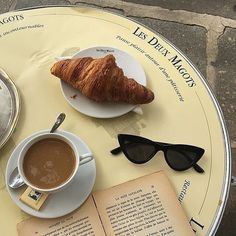 an open book, coffee cup and croissant on a table with reading glasses