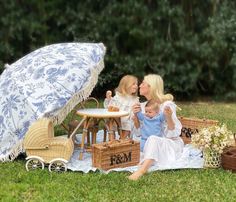 two women and a baby sitting on a blanket in the grass with an umbrella over them