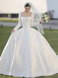 a woman in a white wedding dress and veil walking on the grass with her hands behind her back