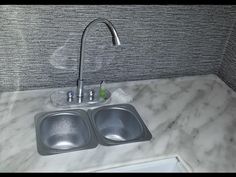 a kitchen sink and faucet with marble counter top in a corner room that has grey tile on the walls