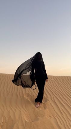 a woman in black is walking across the sand dunes with her veil blowing in the wind