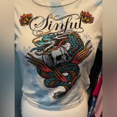 This Sinful Dragon Fire Skull Cropped T-Shirt Features A Bold, Eye-Catching Design With Rhinestones And A Dragon Graphic That Stands Out. The Cameron Wash Gives It A Unique Look, And The Cinch Tie Sides Allow For A Customizable Fit. Made From 100% Cotton, It's Comfortable For Everyday Wear. Brand New With Tags, Never Worn, This Lightweight, Breathable Shirt Is Perfect For Edgy, Casual Style. It Pairs Easily With Just About Anything, Making It An Effortless Addition To Any Wardrobe. Skull Print Crew Neck Top For Festival, Crew Neck Skull Print Top For Festivals, Cotton Tops With Skull Print For Festivals, Edgy Casual Style, Sinful Clothing, Fire Skull, The Cinch, Dragon Graphic, Dragon Fire