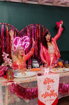 two women dressed in pink are sitting at a table with plates and confetti