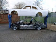 two men working on a car in front of a fenced off area with another man standing next to it