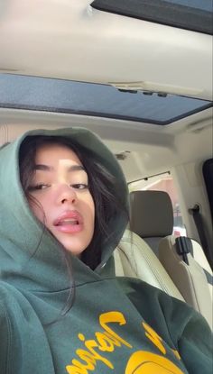 a woman sitting in the back seat of a car wearing a green hoodie and holding her eyes closed