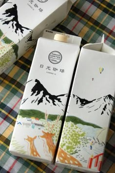 two packages of milk sitting on top of a plaid tablecloth with mountains in the background