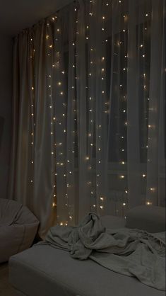 an unmade bed with lights on the curtains