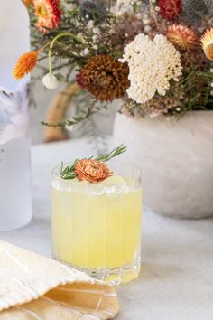 a yellow drink sitting on top of a white table next to flowers and napkins