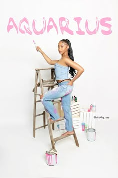 a woman sitting on top of a ladder painting the word aquarius with pink paint