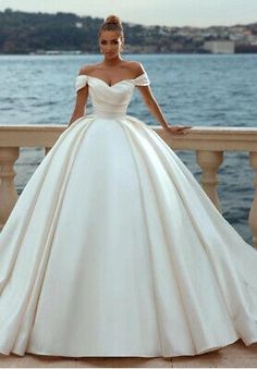 a woman in a white wedding dress standing on a balcony near the water with her hands on her hips