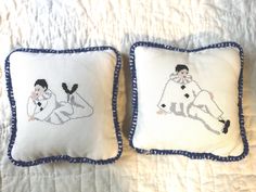 two embroidered pillows on a bed with white sheets and blue trimmings, one is holding a woman's head