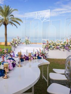 a white table with flowers and candles on it next to some chairs in front of the ocean