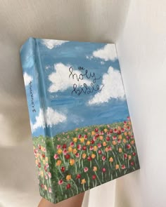 a hand holding up a book with the words happy birthday written on it in front of a field of flowers