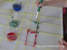 two children are painting squares with red, green and blue paint on paper while another child is holding a brush