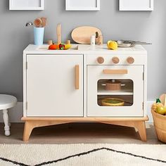 a white wooden play kitchen with pictures on the wall above it and fruit in a basket next to it