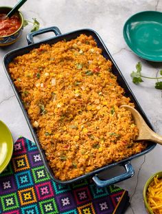 a casserole dish filled with rice and vegetables on a colorful place mat next to bowls of sauces