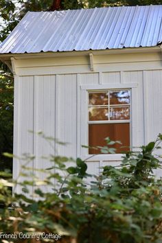 a small white building with a metal roof and window on the side, surrounded by trees