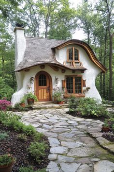 a small white house in the middle of some trees and bushes with potted plants