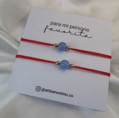 two red string bracelets with blue beads and gold bead on white fabric background