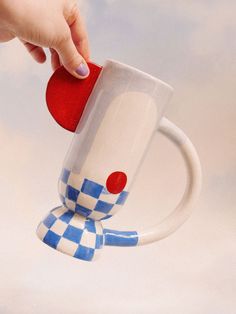 a hand holding a coffee mug with a red ball in it's center and a blue and white checkerboard design on the bottom