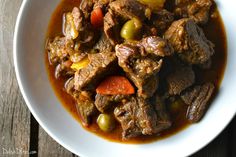 a white bowl filled with meat and vegetables on top of a wooden table next to a spoon