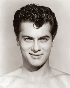 black and white photograph of a man with curly hair smiling at the camera, wearing no shirt