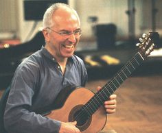 a man sitting on the floor holding a guitar and smiling at the camera with his right hand